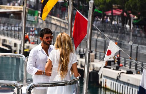 Man and woman stand on bow of a luxury yacht having a chat, with race circuit visible in background
